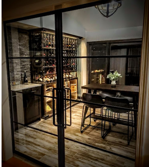 A cozy glass-enclosed wine room with wall-mounted bottle racks, a rustic dining table, and warm ambient lighting