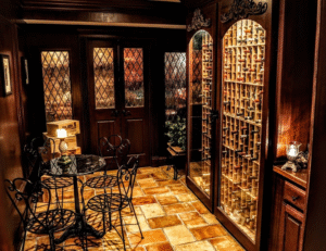 A cozy, warmly lit wine cellar with wooden racks full of bottles, tiled floors, and a small wrought-iron table and chairs