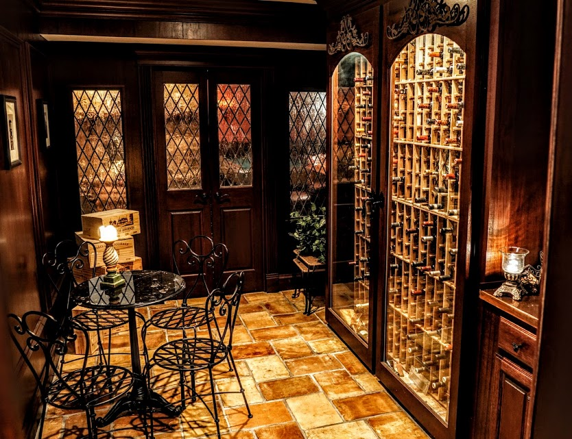 A cozy, warmly lit wine cellar with wooden racks full of bottles, tiled floors, and a small wrought-iron table and chairs