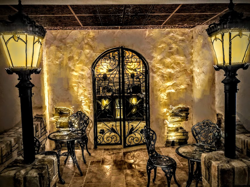 An elegant stone wine cellar entrance with wrought-iron doors, warm lighting, and small café-style tables and chairs